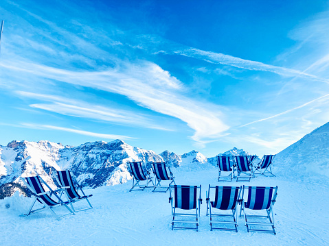 Breathtaking peaks with the beauty and majesty of the Alps. Craggy snow-covered peaks in the stunning Alps and a breath of pure mountain air. Empty blue deck chairs on the snow in ski resort, Austrian Alps.