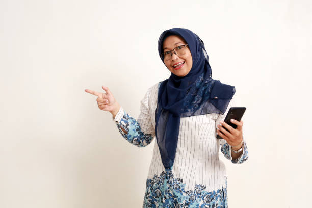 istock Asian elderly muslim woman standing holding a cell phone while pointing sideways 1984870026