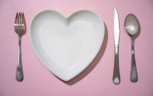 Heart plate with silverware of fork, knife, and spoon on pink background. Romantic dinner Valentine's Day concept