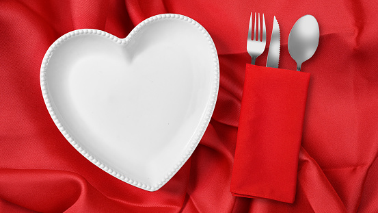 Heart plate with silverware of fork, knife, and spoon in red napkin over a red napkin background. Romantic dinner Valentine's Day concept