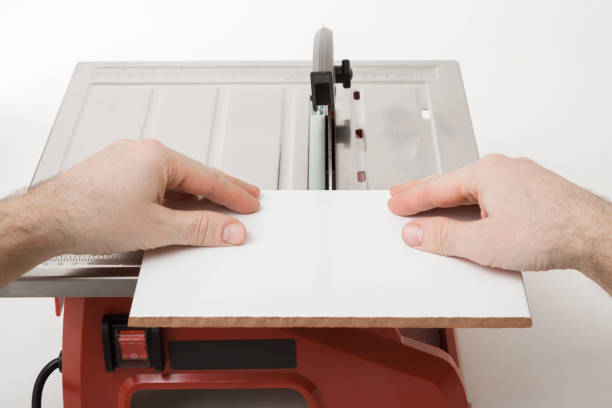 Young adult man hand using electrical saw and cutting new white ceramic tile on light gray table background. Preparing for tiles laying. Closeup. Renovation process. Point of view shot.