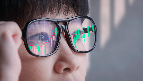 Asian Contemplative female Crypto Trader Looking at computer screen while Analyzing online Stock Trading Data Chart with light Reflection of stock Graph Chart on Eyeglasses Surface