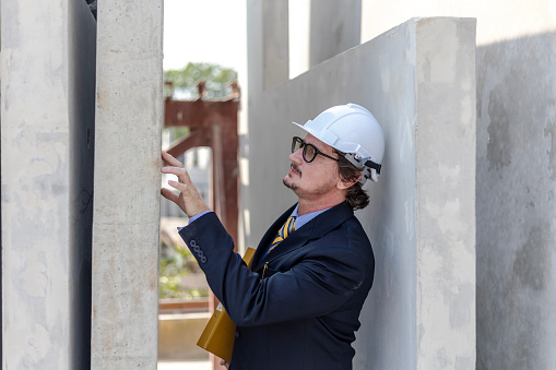 Male engineer checks quality of readymade floor or wall in a factory. Technician with safety hat stands next to a pile of concrete walls.