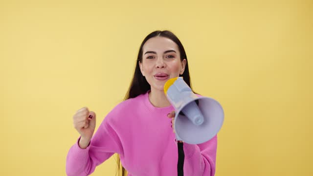 Cheerful female holding loudspeaker and telling announcement while making win gesture. Smiling woman talking in megaphone and supporting friends while standing in studio with yellow background.