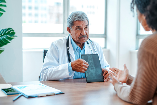 Senior male physician with mustache, wearing lab coat with stethoscope around neck holding up digital tablet showing spinal x-ray, explaining result to female patient in clinic