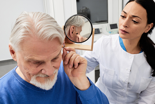 Senior man trying on hearing aid in front of mirror with female audiologist, fitting hearing device into his ear while audiology consultation