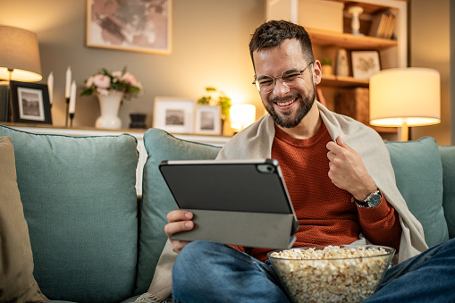 A smiling young man is relaxing at home eating popcorn and watching a movie online on his favorite tv platform