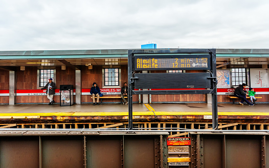 Boston, Massachusetts, USA - February 1, 2024: People waiting on the platform of an outdoor train station. Massachusetts Bay Transportation Authoritys (MBTA) Red Line subway station in the Beacon Hill neighborhood of Boston. The very top of the distant 200 Clarendon Street skyscaper (aka John Hancock Tower)  is visible in the background.