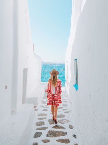 Back view of a young beautiful happy traveler female lady with a red dress enjoys the scenery and walking through the turquoise-colored Mediterranean seawater from the traditional white and blue small, whitewashed alleys of Mikonos town, Cyclades islands of Greece during summer time on windy and sunny day