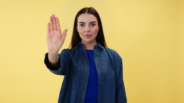 Caucasian woman doing stop sign with palm while looking seriously at camera. Young lady standing in studio with yellow background while expressing disapproval and rejection of something unpleasant.