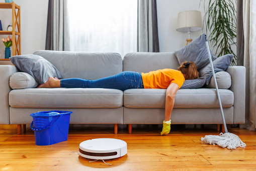 An exhausted individual lies on a couch with a mop, with a robot vacuum cleaner and bucket in the foreground.