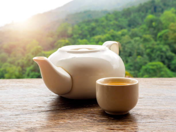 istock Teapots set. White ceramic teapot with hot tea water in small cup preparing on wooden table on natural green mountain view, countryside background with sunlight in the morning. 1985229019