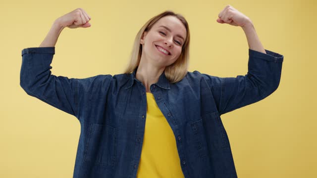 Sporty young woman looking at arms muscles and smiling while standing in studio with yellow background. Athletic female showing biceps and feeling proud and confident in powers for achieving goals.
