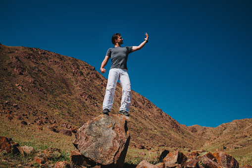 Handsome young man stands on big stone on mountains background and looks at his hand