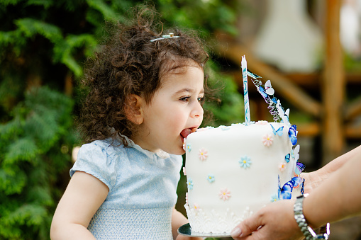 Two years old toddler girl with curly hair biting her birthday cake in the garden with vibrant spring colours. Outdoor vibrant colours