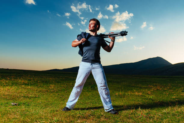 Handsome young man photographer with tripod posing on sky and mountains background at twilight