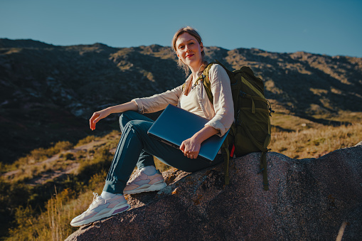 Young woman tourist with backpack and laptop sits on top of the mountain