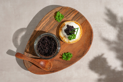 Black caviar on mini blini or pancake with glass jar on a wooden plate, top view.
