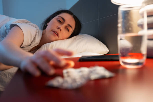 una mujer adulta caucásica alcanza una mesita de noche roja con medicamentos. mano desenfocada y pastillas para dormir en primer plano. concepto de insomnio, trastornos del sueño y dolor de cabeza - narcotic medicine pill insomnia fotografías e imágenes de stock