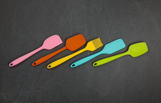 Five silicone cooking spatulas and brushes on black slate board with copy space, colorful modern culinary kitchen utensils, close up, elevated table top view, directly above