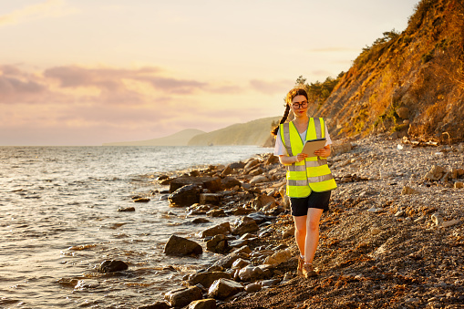 Inspection environmentalist. Volunteer woman wearing vest and using pad for analyzing see water. In background is ocean, coast and sunset. Concept of environment pollution.
