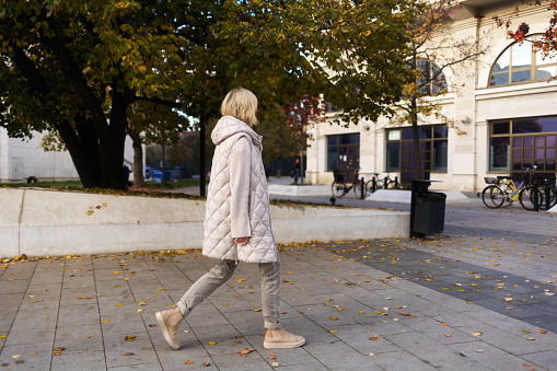 Alone blonde woman wearing white jacket walking at street. Side view. Concept of psychology and loneliss.