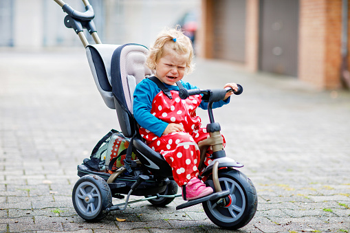 Cute adorable toddler girl sitting on pushing bicycle or tricycle. Little baby child going for a walk with parents cold cloudy day. Happy healthy kid in colorful clothes.