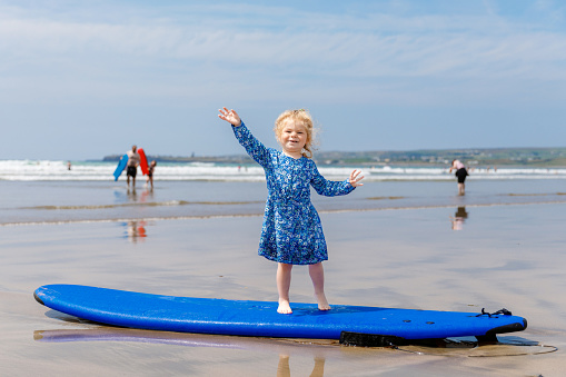 Little cute toddler girl at the Ballybunion surfer beach, having fun on surfboard for the first time, west coast of Ireland. Happy child enjoying Irish summer with family