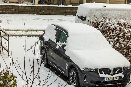 Sweden. Uppsala. 02.04.2024. View of electric vehicle BMW iX3 blanketed in snow, connected to charging cable, in parking lot private residence on wintry day.