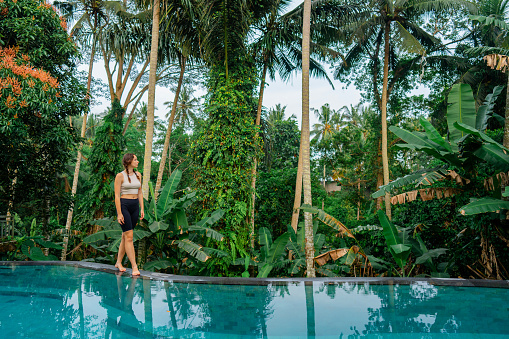 Woman walking on the edge of the swimming pool  in the morning, she is surrounded by lush green  jungles
