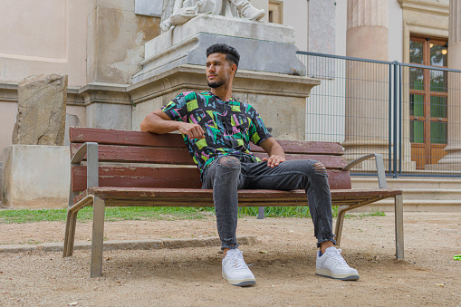 a nice style moroccan man sitting on a wooden bench of the street while looking aside | full length