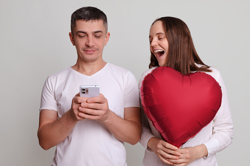 Man and laughing woman wearing white clothing holding heart shaped air balloon isolated over gray background using cell phone browsing internet web pages