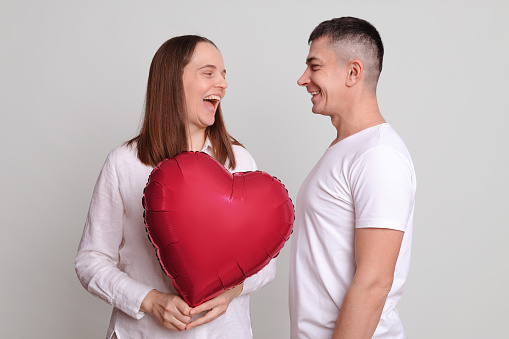 Laughing woman and man wearing white clothing holding heart shaped air balloon looking at each other expressing positive emotions satisfied couple isolated over gray background