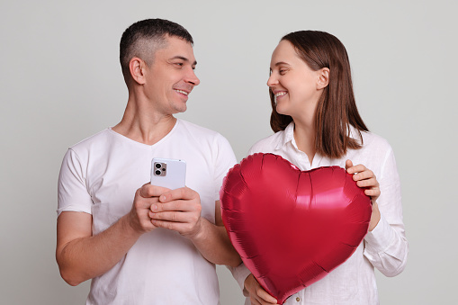 Cheerful attractive man and woman wearing white clothing holding heart shaped air balloon using mobile phone scrolling online with happy facial expressions isolated over gray background