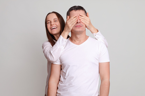 Excited young couple guy and girl posing isolated over gray background wife covering husband eyes with hands having fun together enjoying happy moments spending together