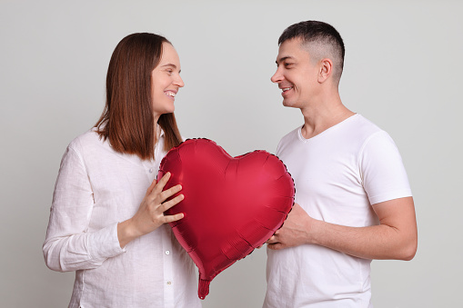 Happy romantic couple enjoying dating man and woman with heart shaped balloon isolated over grey background looking at each other with love and gentle