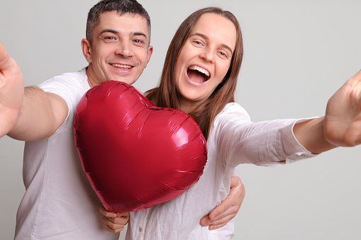 Overjoyed cheerful couple man and woman with heart shaped balloon isolated over grey background making point of view photo laughing happily while celebrating St. Valentines Day