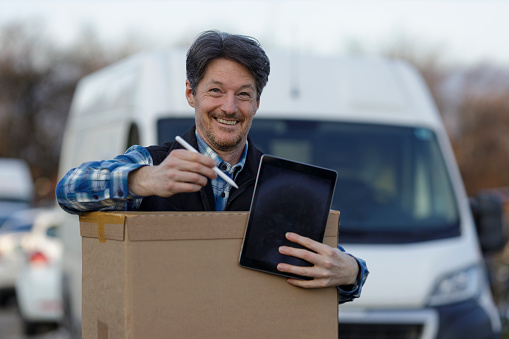 Cheerful courier holding a big cardboard box while standing in front of his van waiting for castumer signature. Menges, Slovenia