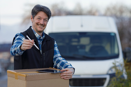 Cheerful courier holding a big cardboard box while standing in front of his van waiting for castumer signature. Menges, Slovenia