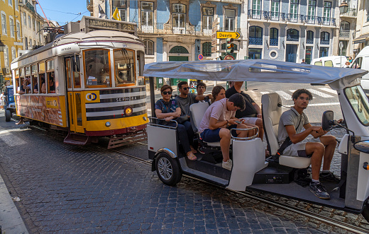 Tourist tuk tuk or three-wheeled Autorickshaw packed with tourists going up a cobblestone street in the Alfama neighborhood of Lisbon, followed by a yellow tram.