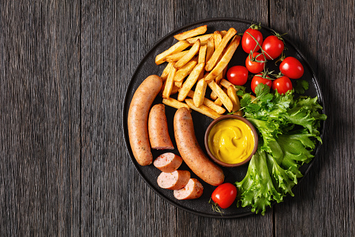 italian sausages with french fries, lettuce, fresh tomatoes and mustard on black plate on dark wooden table, horizontal view from above, flat lay, free space