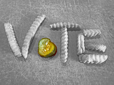The word VOTE spelled out on a wooden cutting board.    Letters V, T, and E spelled out with french fries.   The letter O is a pickle.  Black and white photograph.  Pickle in color.