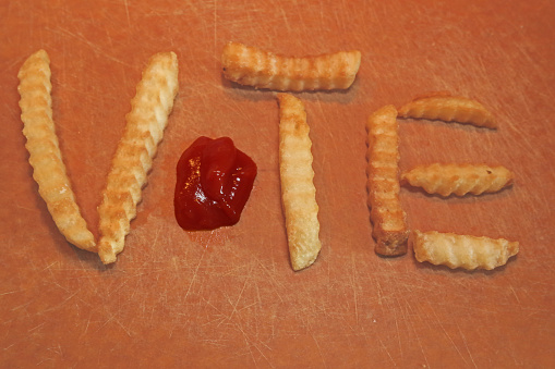 The word VOTE spelled out on a wooden cutting board.    Letters V, T, and E spelled out with french fries.   The letter O ketchup.