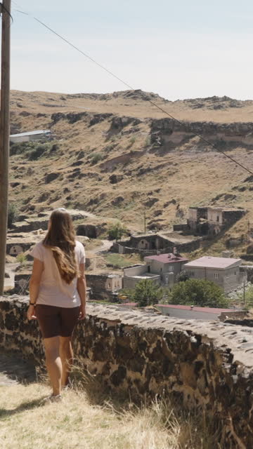 Vertical video. A young woman walks through the ancient town of Saratli in Turkey, with an overhead view of houses at the base of the hills.