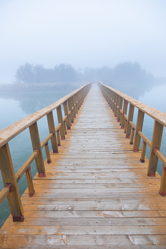Vertical image of a quiet wooden footbridge leads to a fog-shrouded islet over calm waters
