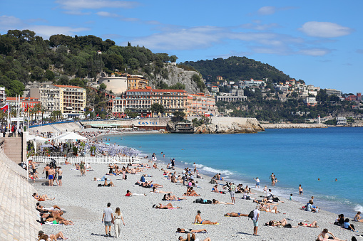 Nice, France - May 2, 2023: The architecture of the city's coastline and sunbathers relaxing on a pebbled Mediterranean beach. It is one of many beaches on a seafront in the city.