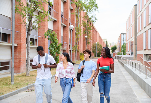 Cheerful multiethnic teenage students walking down the street with notebooks and looking at each other while talking against the brick buildings on the university campus. Concept of education and friendship