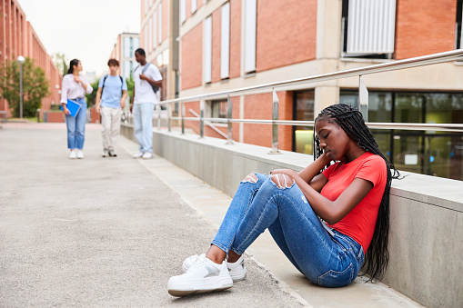 Sad young African American student girl as her friends talk in the background having fun on the college campus. Bullying at school. Concept of adolescent mental health