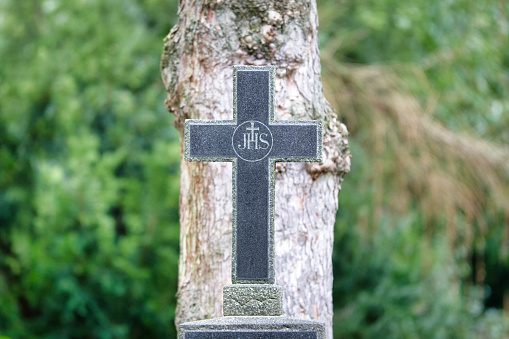 The Christian IHS monogram on a stone cross in a cemetery in front of a tree in a blurred background