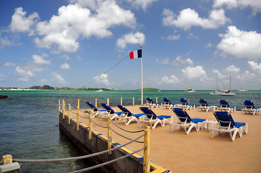 Marigot, Collectivity of Saint Martin, French Caribbean: pier with deckchairs and the French flag - pier extending into the tropical clear turquoise waters of a Marigot bay beach, surrounded by anchored sailboats.